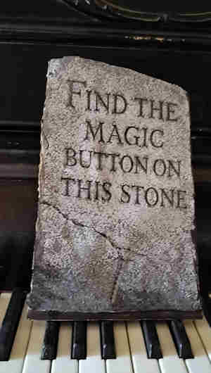 A printed paper tombstone standing upright on a piano keyboard, reading "Find the magic button on this stone"